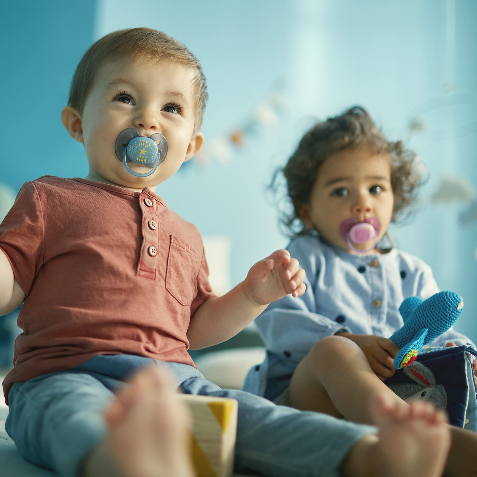 Two toddlers sitting on play mat with toys one using Philips Avent ultra soft pacifier UAE blue and another with pink pacifier