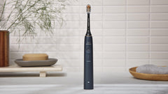 Philips Sonicare 9900 Prestige toothbrush matte black standing on bathroom counter with soap dish towel and tiled white wall
