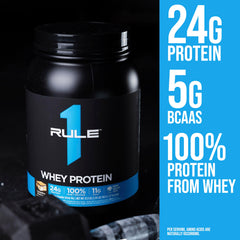 Black Rule 1 whey protein tub labeled Rule1 1 Whey Blend 24g protein next to chocolate pieces and shaker bottle
