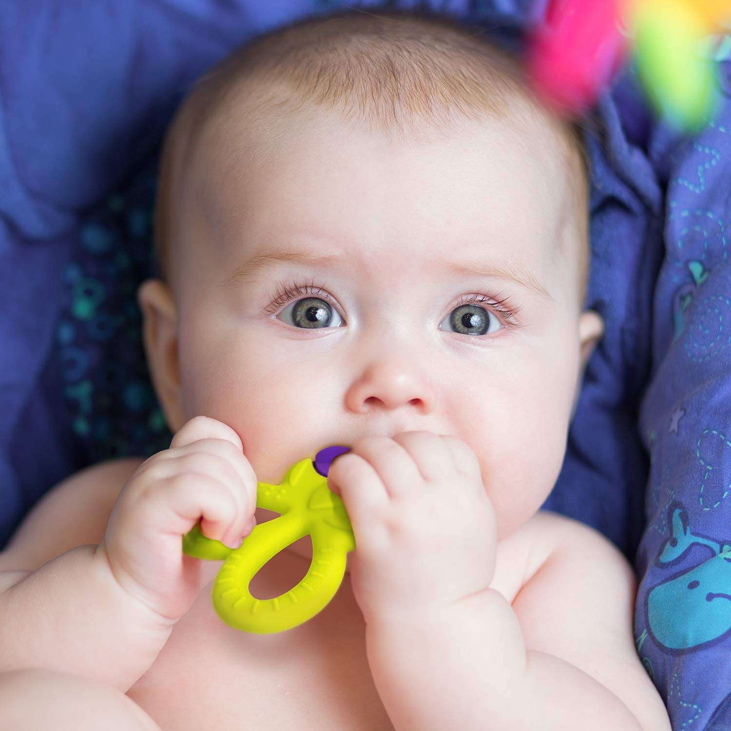 Infant chewing bright green silicone baby teether UAE while seated in a blue patterned baby seat
