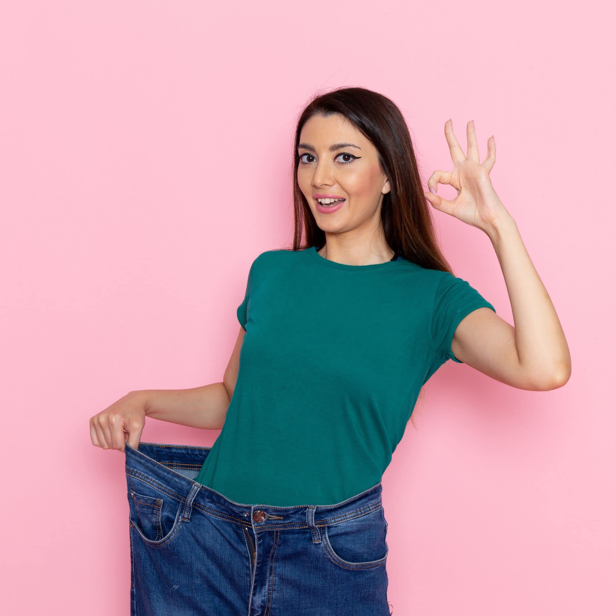 Woman in oversized jeans holding waistband and OK sign on pink background with saffron slimming tea for weight loss
