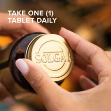 Hand holding a single round white tablet with SOLGAR imprint and bottle behind