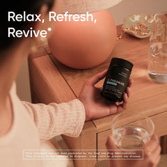 Hand holding Sports Research L-theanine softgels bottle by bedside table with glass of water and lamp