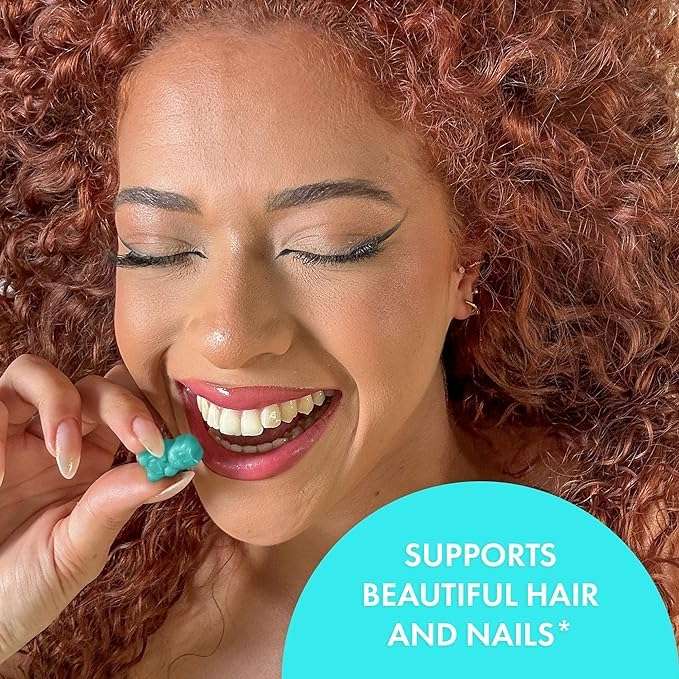 Smiling woman with curly red hair holding a teal SugarBearHair vegan hair vitamins gummies piece