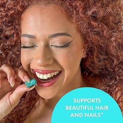Smiling woman with curly red hair holding a teal SugarBearHair vegan hair vitamins gummies piece