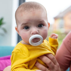 Baby in yellow outfit held by caregiver using white Tommee Tippee breast-like pacifier closeup in home setting