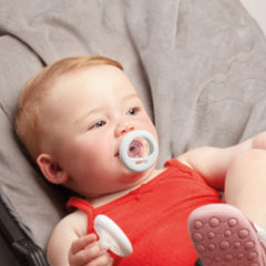 Baby in red onesie lying on gray blanket using a Tommee Tippee breast-like pacifier for soothing