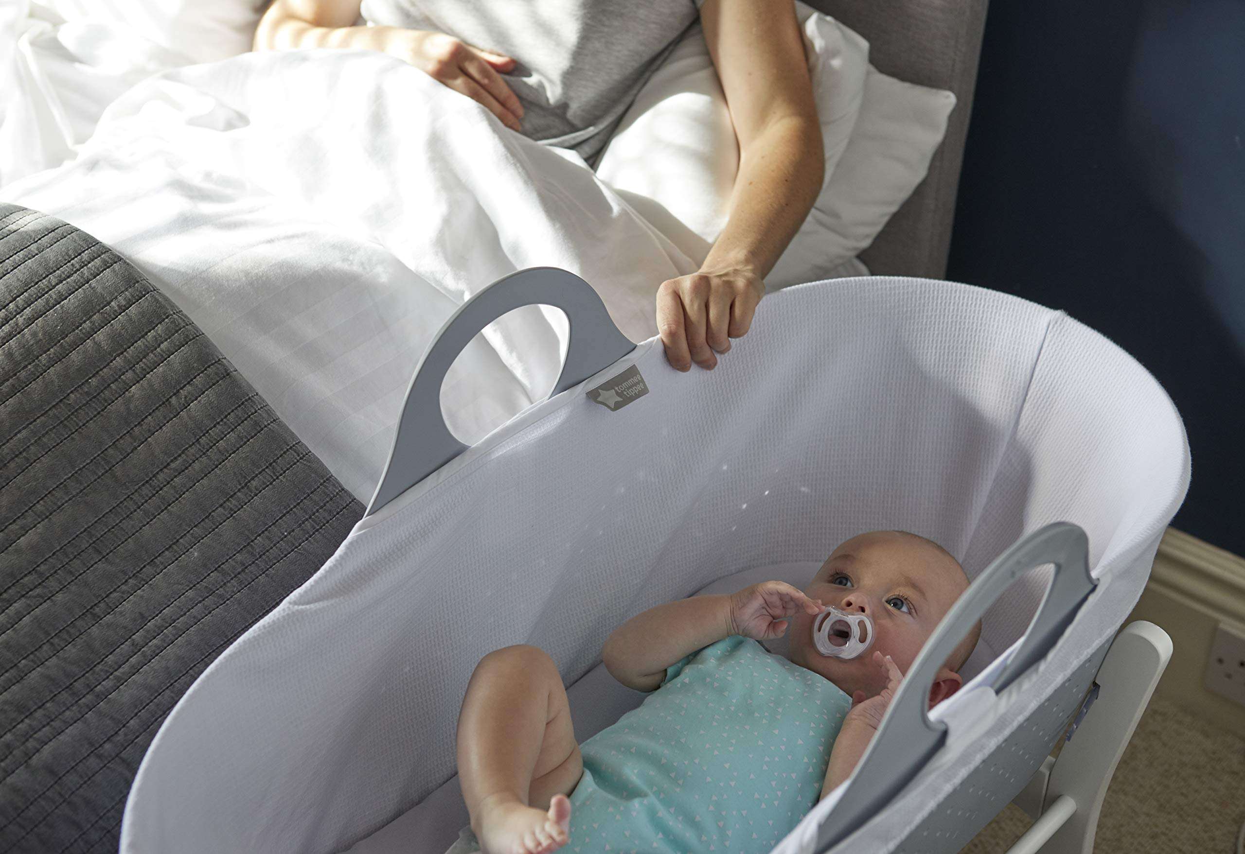 Baby in white bedside bassinet using Tommee Tippee ultra light soother while parent reaches from bed