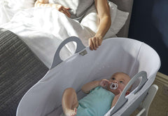 Baby in white bedside bassinet using Tommee Tippee ultra light soother while parent reaches from bed