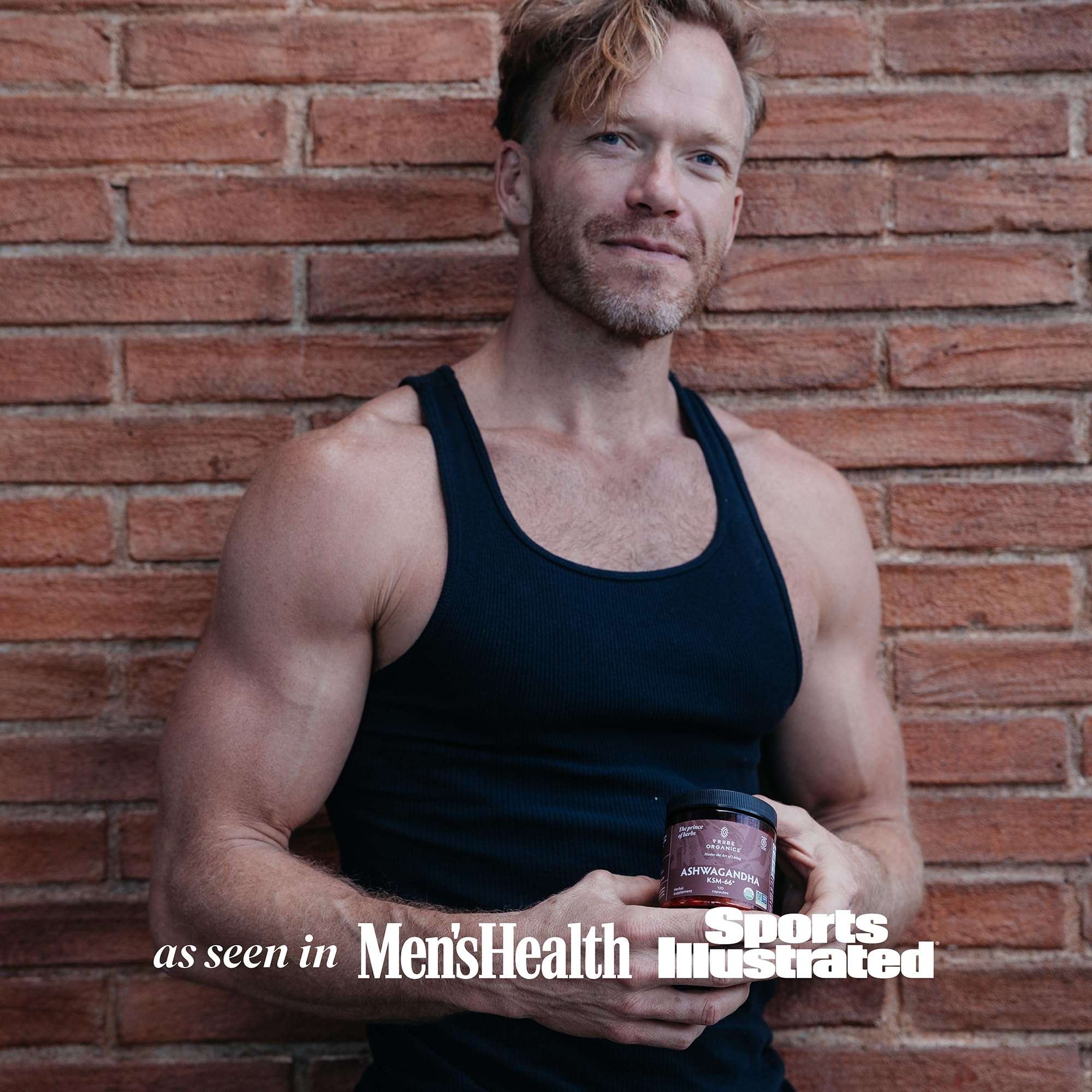 Muscular man in black tank top holding TRIBE ORGANICS jar of Ashwagandha KSM-66 capsules UAE against brick wall