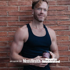 Muscular man in black tank top holding TRIBE ORGANICS jar of Ashwagandha KSM-66 capsules UAE against brick wall