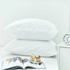 Two white cotton bed pillows UAE stacked on a bed next to a bedside table with a clock and an open magazine