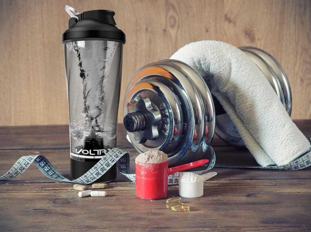VOLTRX electric protein shaker bottle with clear body and mixing vortex beside dumbbell towel and scoops