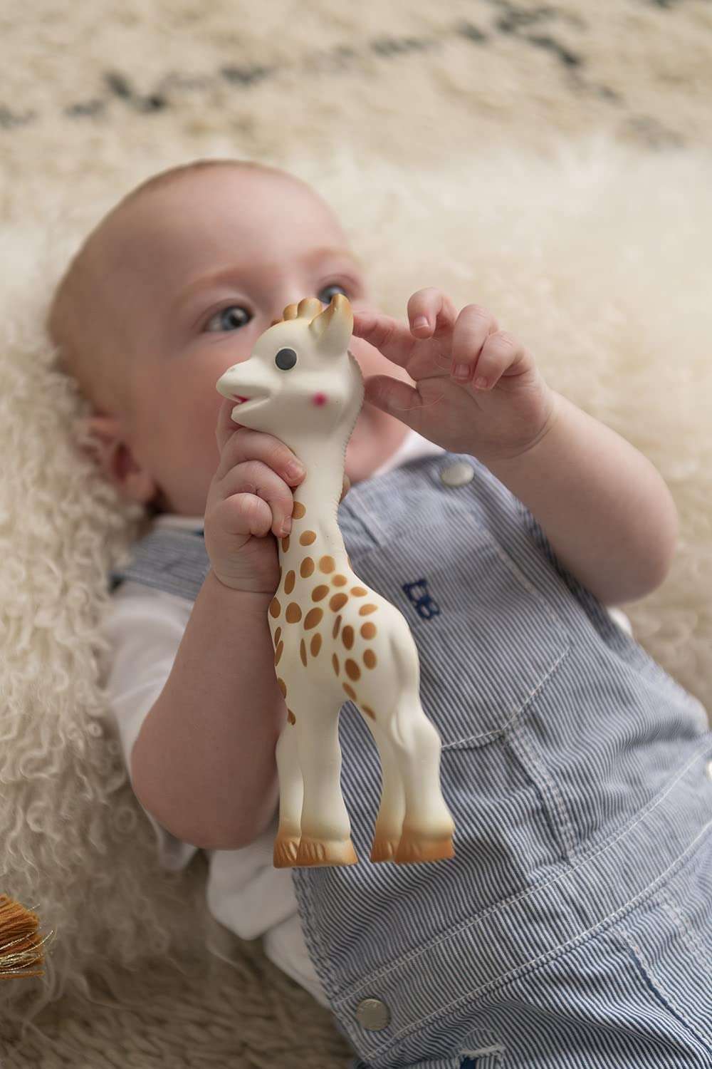 Baby lying on soft rug holding Sophie La Girafe teether UAE chew toy in blue striped overalls for soothing