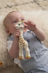 Baby lying on soft rug holding Sophie La Girafe teether UAE chew toy in blue striped overalls for soothing