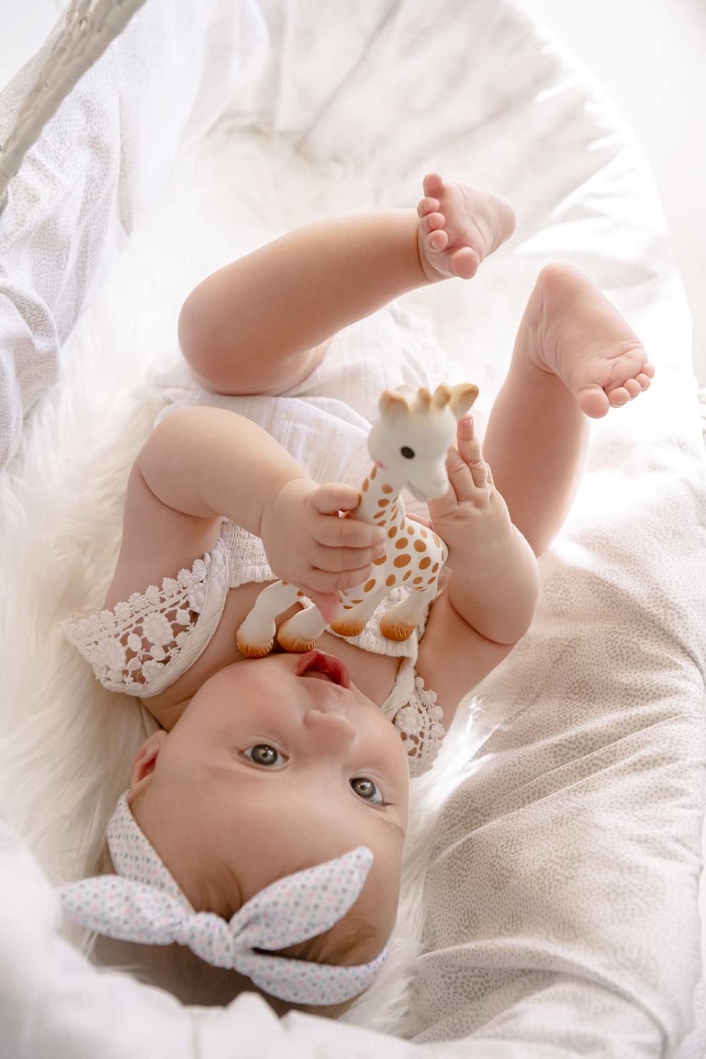Baby in white bassinet holding Sophie La Girafe teether UAE toy wearing lace outfit and headband