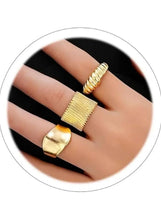 Close-up of gold-tone rings on hand: textured open band, wide cuff, and ridged ring