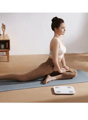 Xiaomi body composition smart scale in white next to a woman practicing yoga on a mat