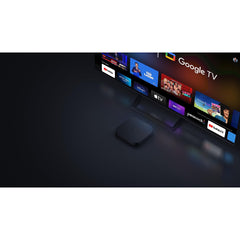 Xiaomi Mi Box S 2nd Gen 4K streaming device placed in front of a TV displaying Google TV interface